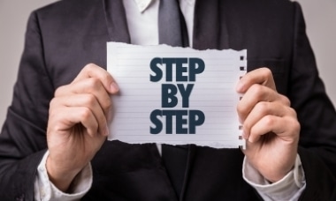 Man in a suit holding a paper that says “STEP BY STEP.”